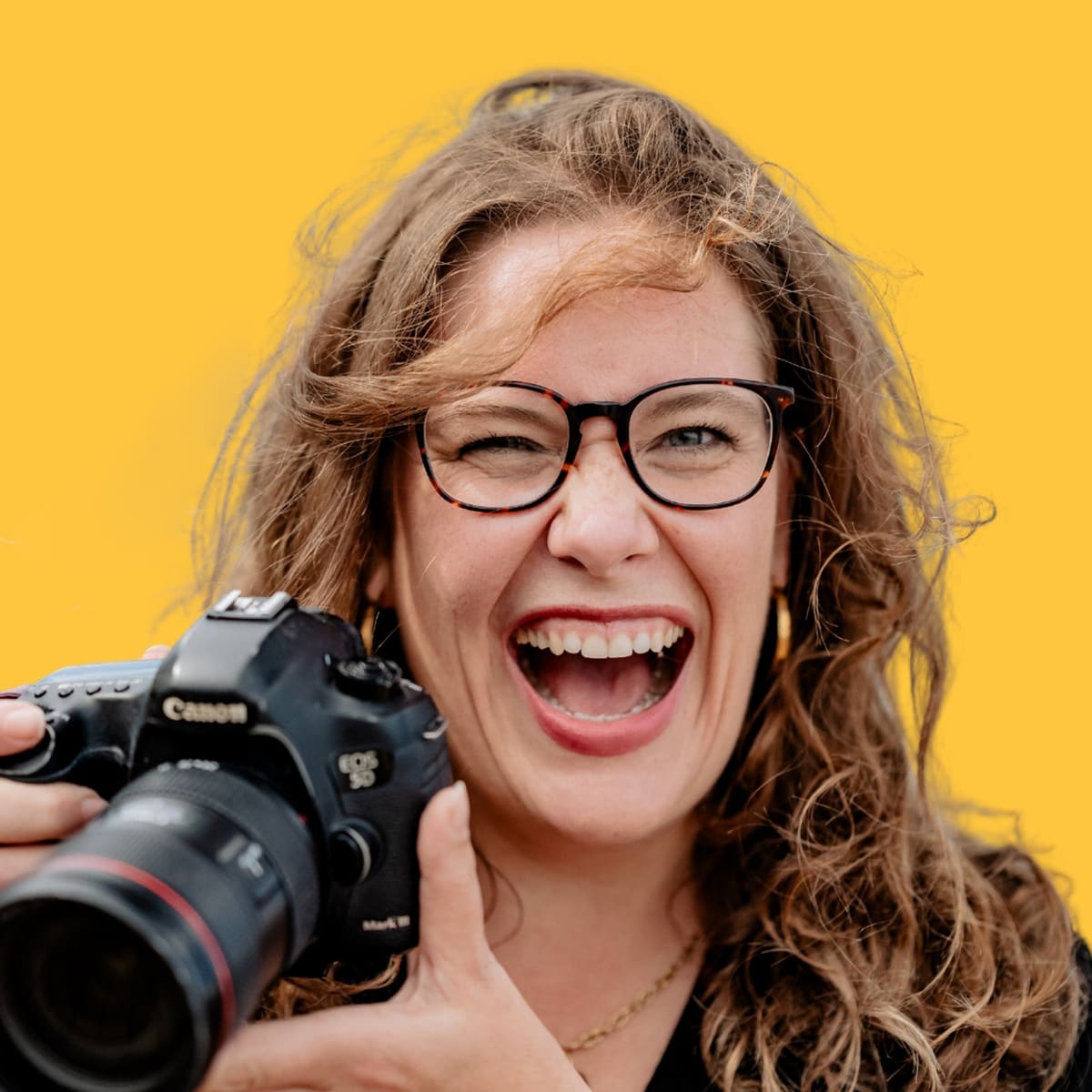 Fotografin Bianca Kübler lacht mit Kamera in der Hand vor gelbem Hintergrund