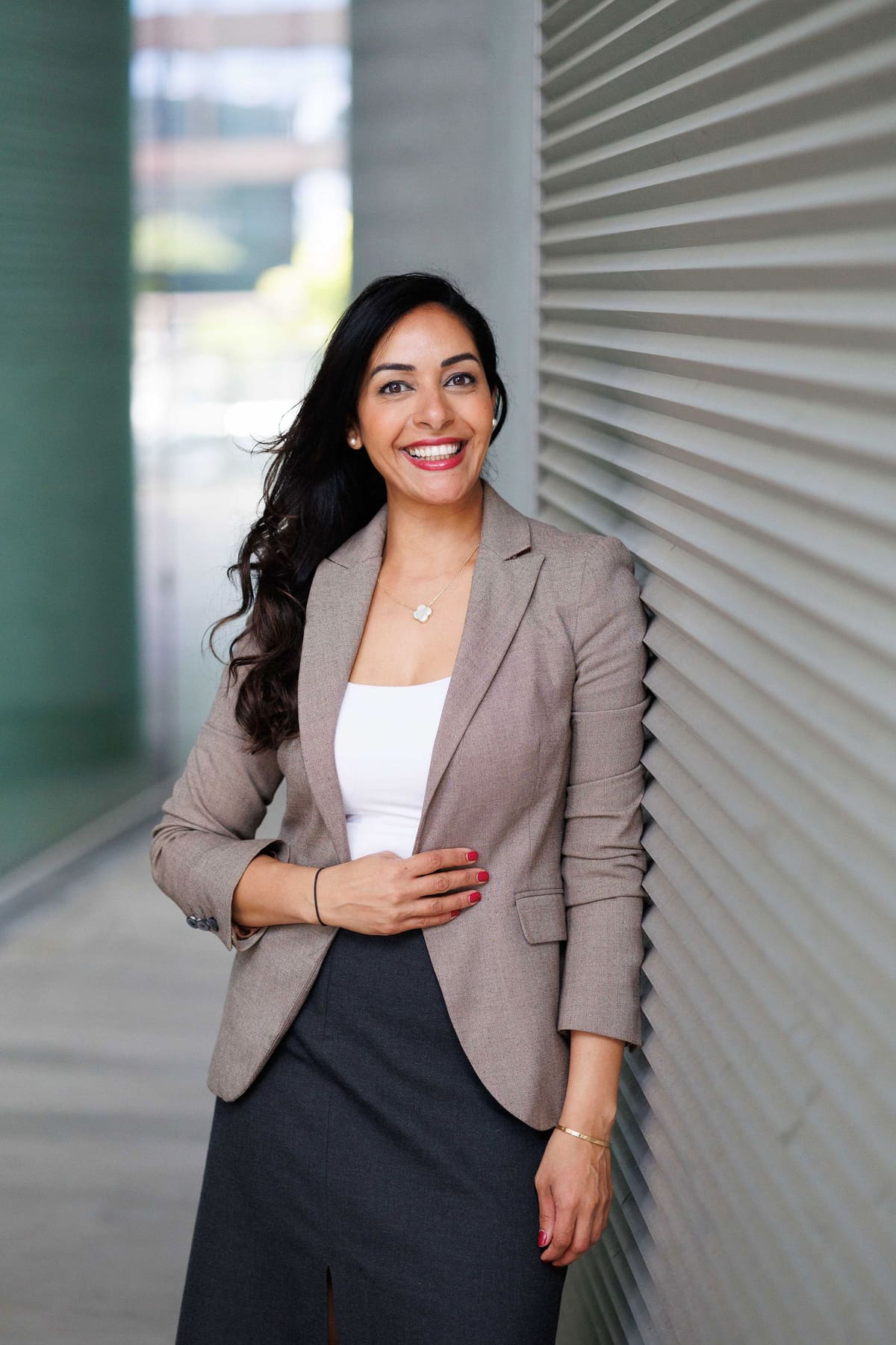 Business-Portrait im Stehen Frau mit langen dunklen Haaren im beigen Blazer lehnt an einer grauen Wand