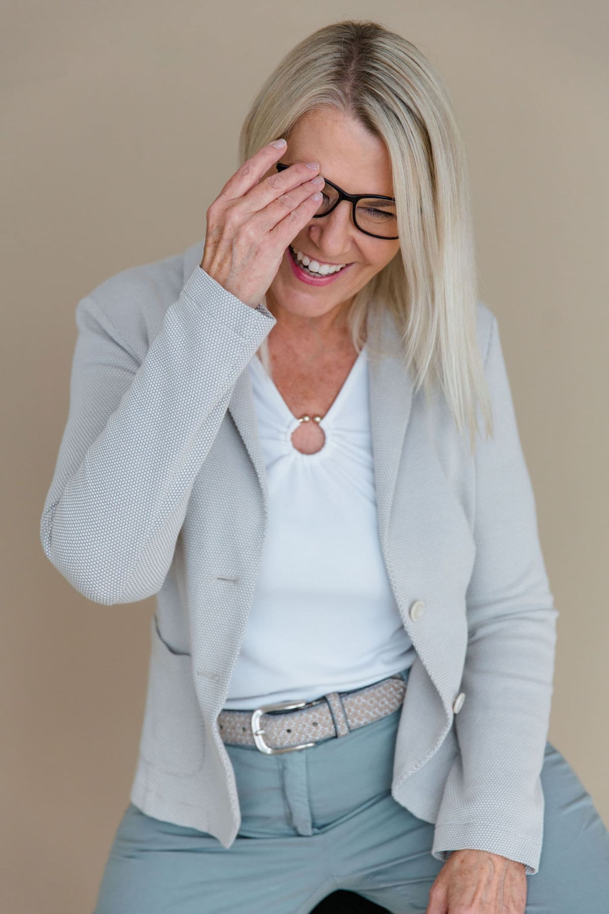 Blonde Frau im grauen Sakko lacht mit der Hand an der Stirn