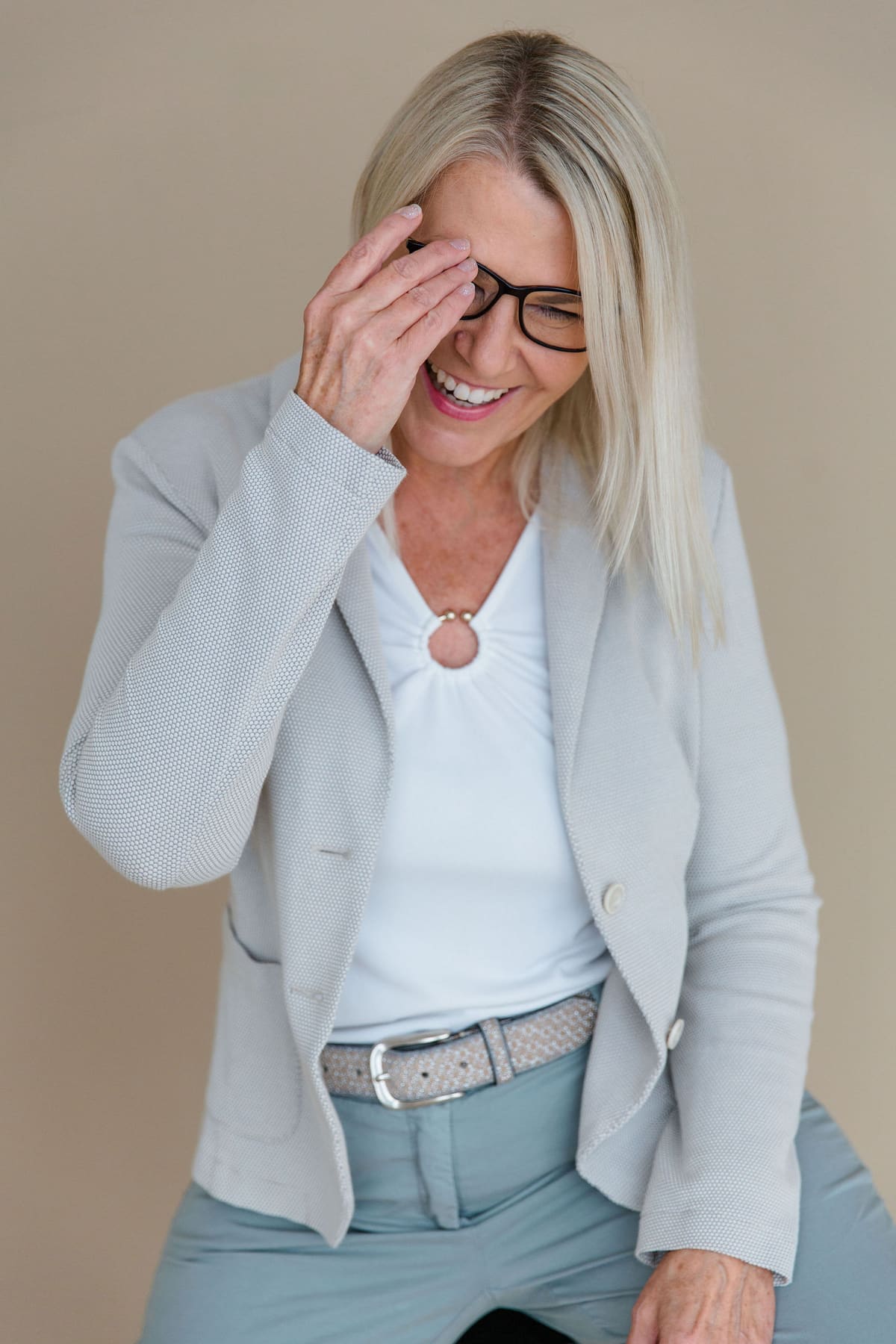 Frau mit blondem Haar, Brille und hellgrauem Blazer lacht und hält sich eine Hand vors Gesicht