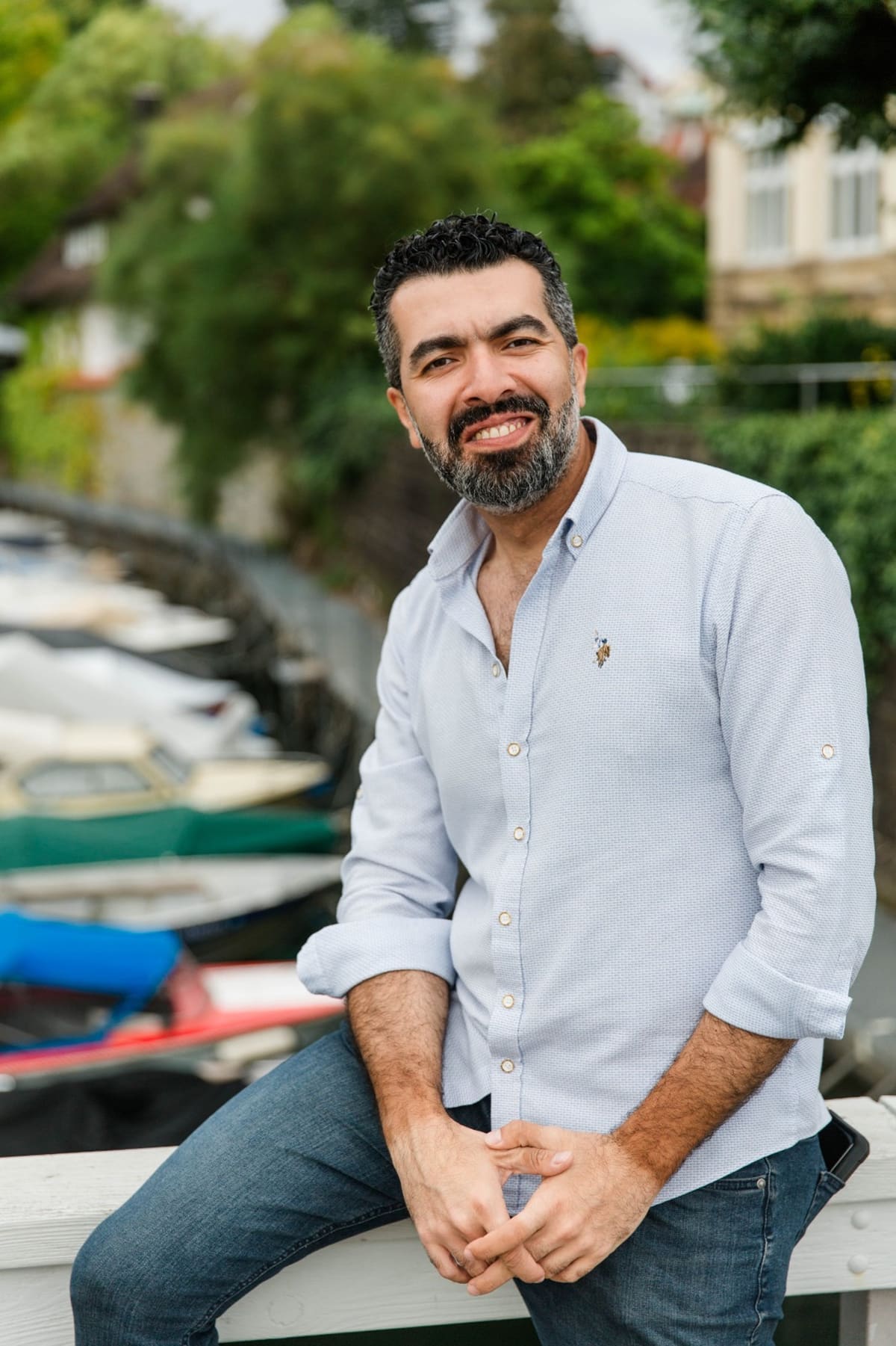 Business-Portrait eines Mannes mit Bart im hellen Hemd, sitzend an einem Ufer mit Booten