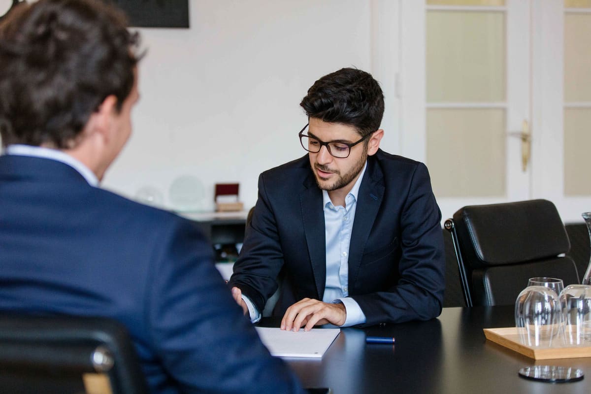 Mann mit Brille im dunklen Anzug schreibt am Besprechungstisch, ein Gesprächspartner von hinten im Vordergrund