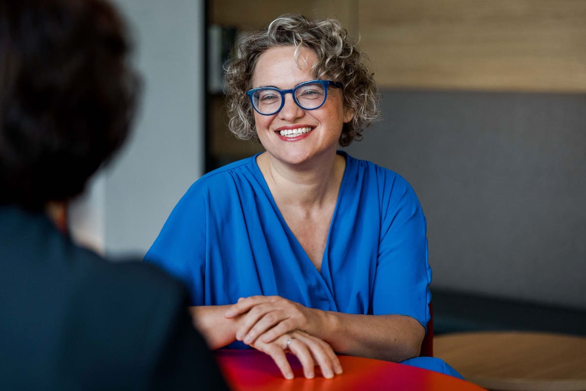 Frau mit Brille und blauer Bluse lacht am Tisch im Gespräch, ihr Gegenüber von hinten