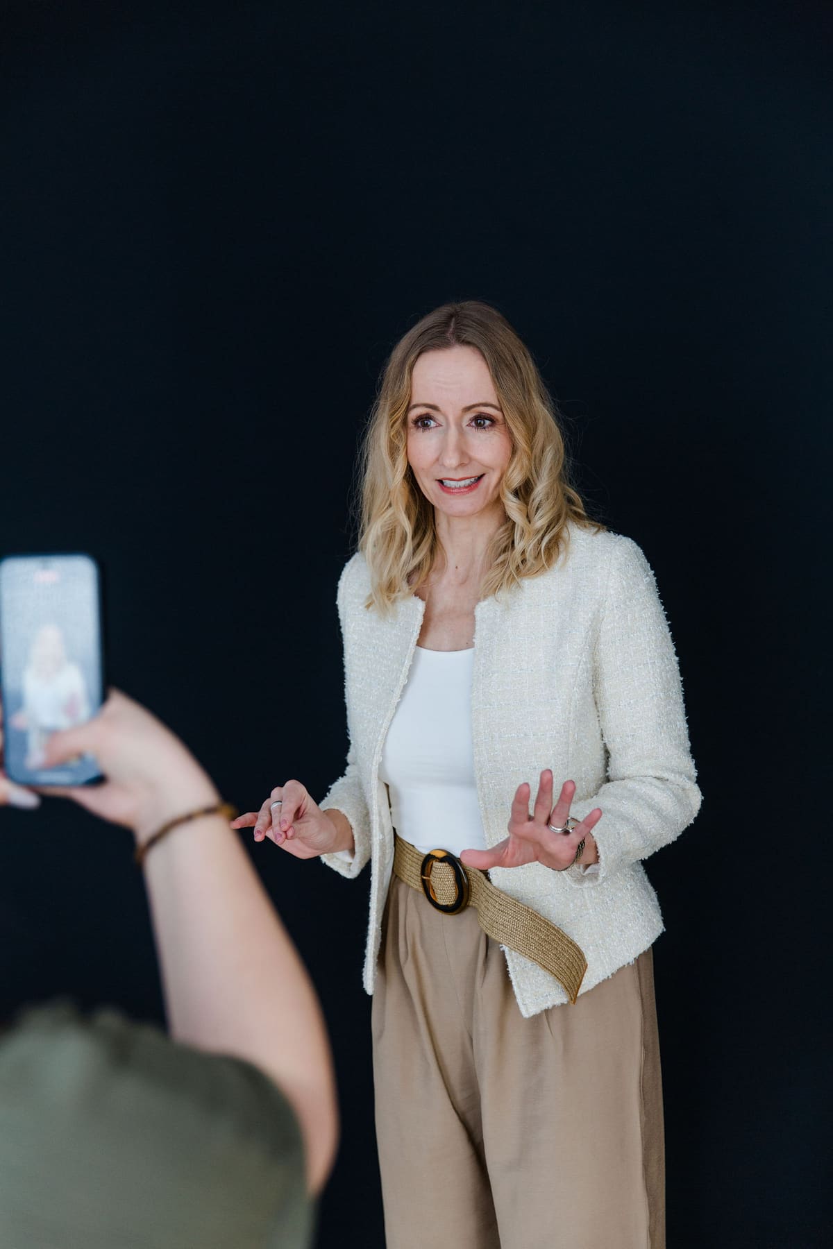 Frau im hellen Blazer spricht vor dunklem Hintergrund, vorne filmt ein Smartphone mit
