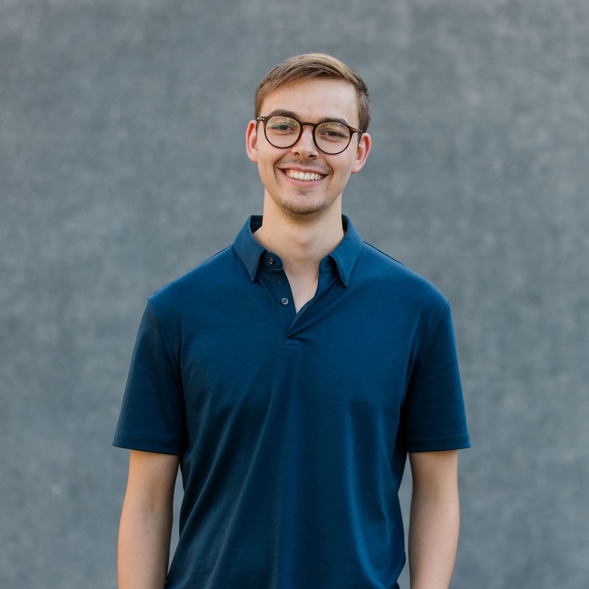Mitarbeiterportrait vor ruhigem Hintergrund Junger Mann mit Brille im dunkelblauen Poloshirt lächelt vor einer grauen Wand