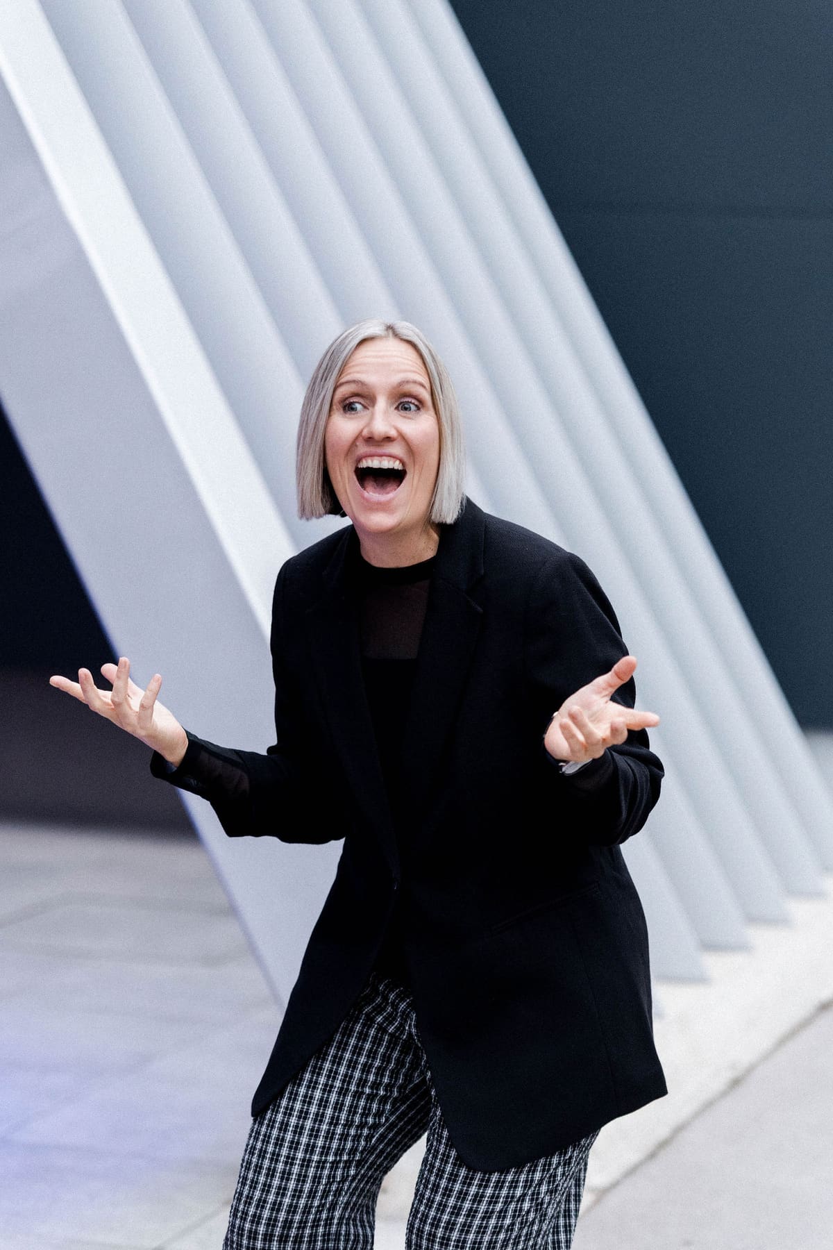 Frau im schwarzen Blazer lacht mit offenen Händen vor heller Architektur
