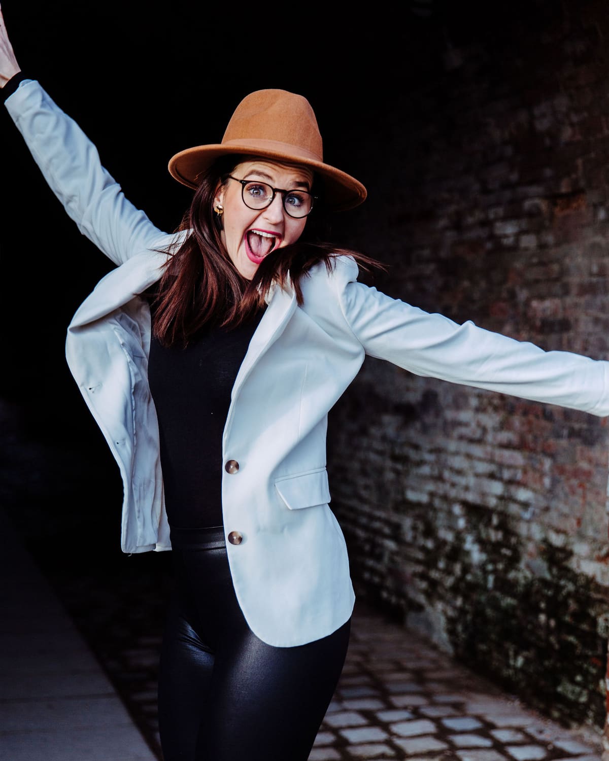 Frau mit Hut und hellem Blazer jubelt mit erhobenem Arm vor einer Ziegelwand