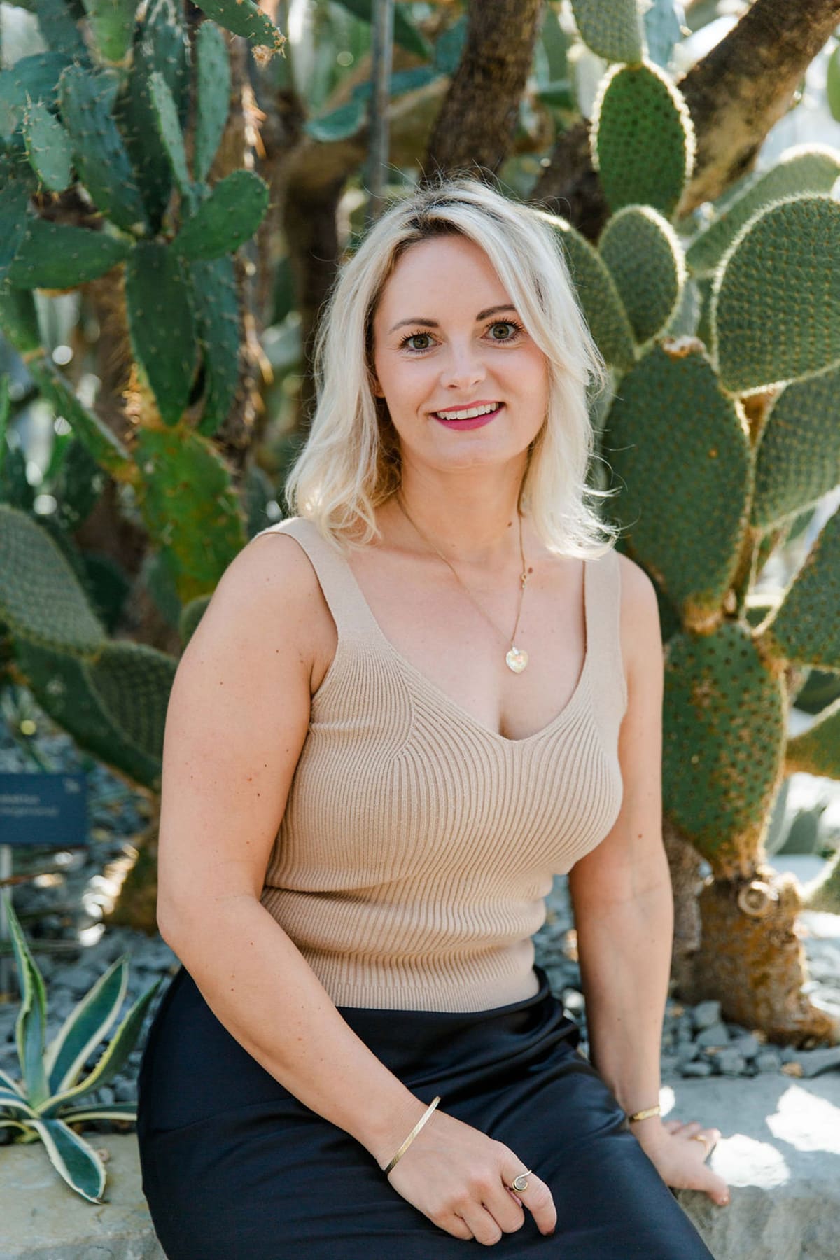 Blonde Frau im beigen Top sitzt lächelnd zwischen großen Kakteen