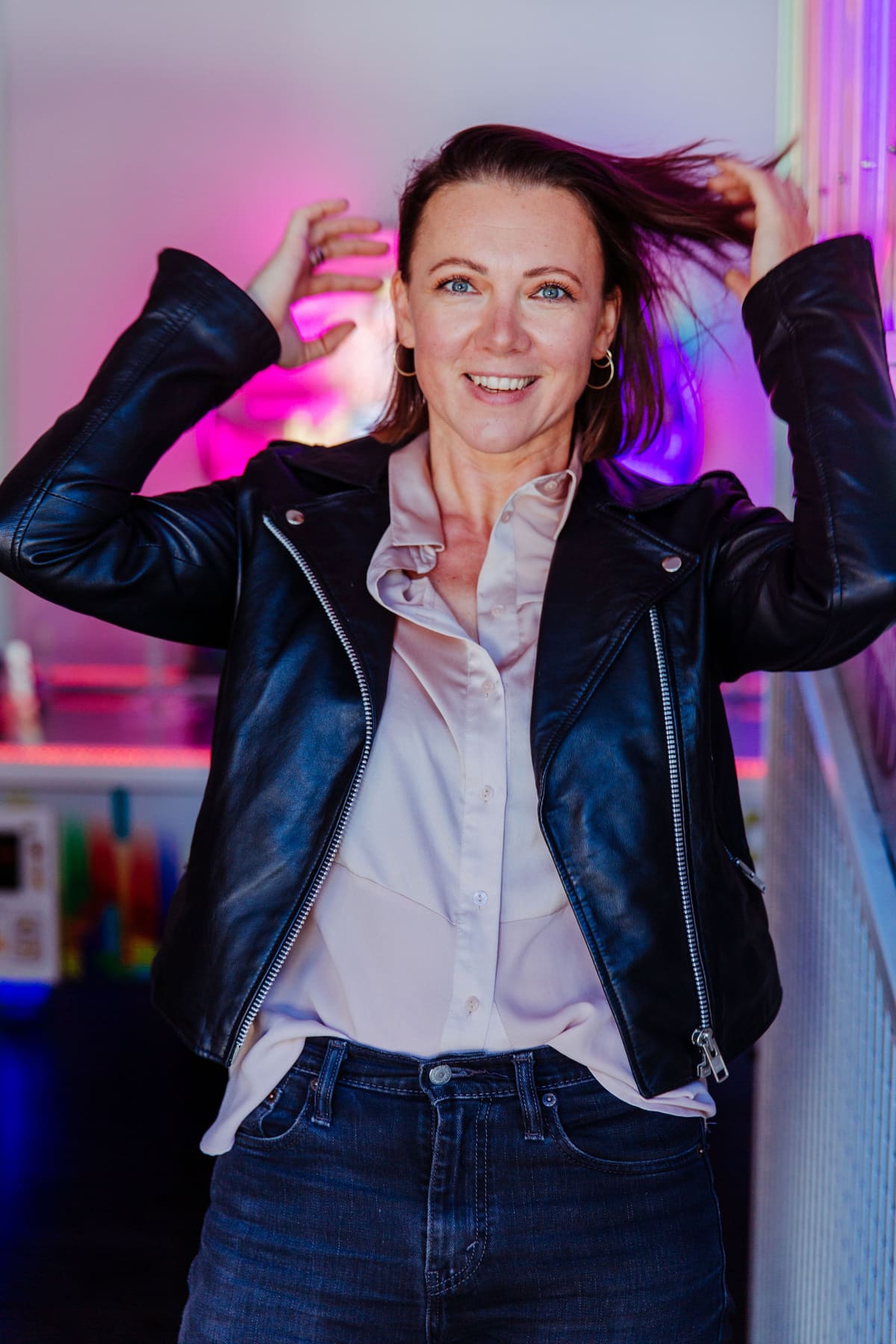 Frau in Lederjacke fährt sich lachend durchs Haar, pinkes Licht im Hintergrund