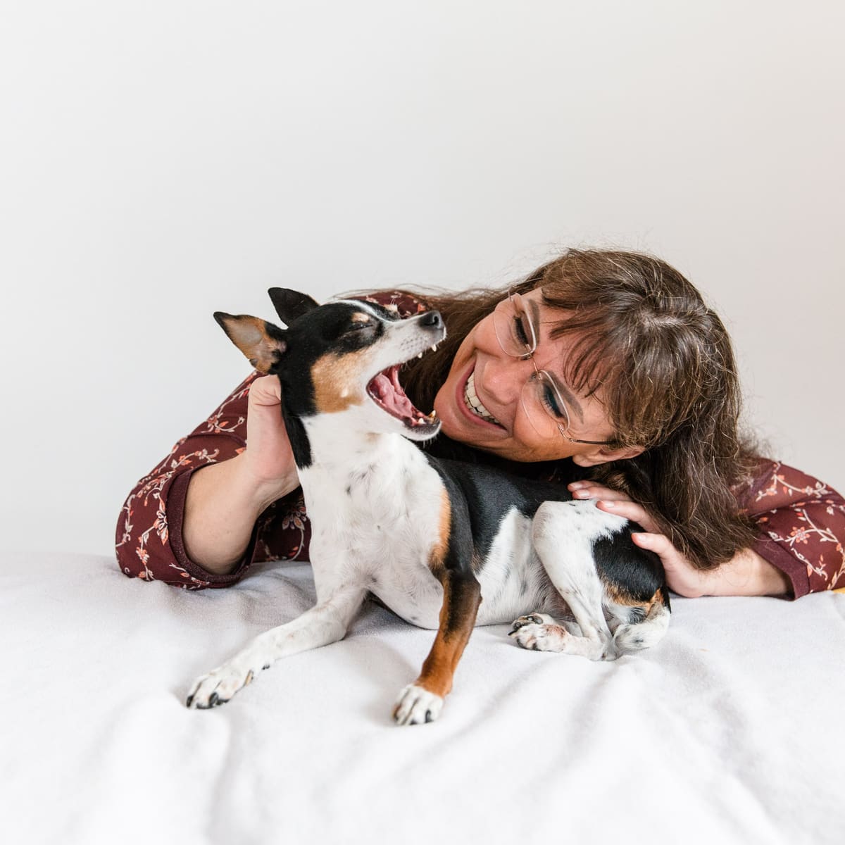 Frau liegt lachend mit ihrem Hund auf einem weißen Bett