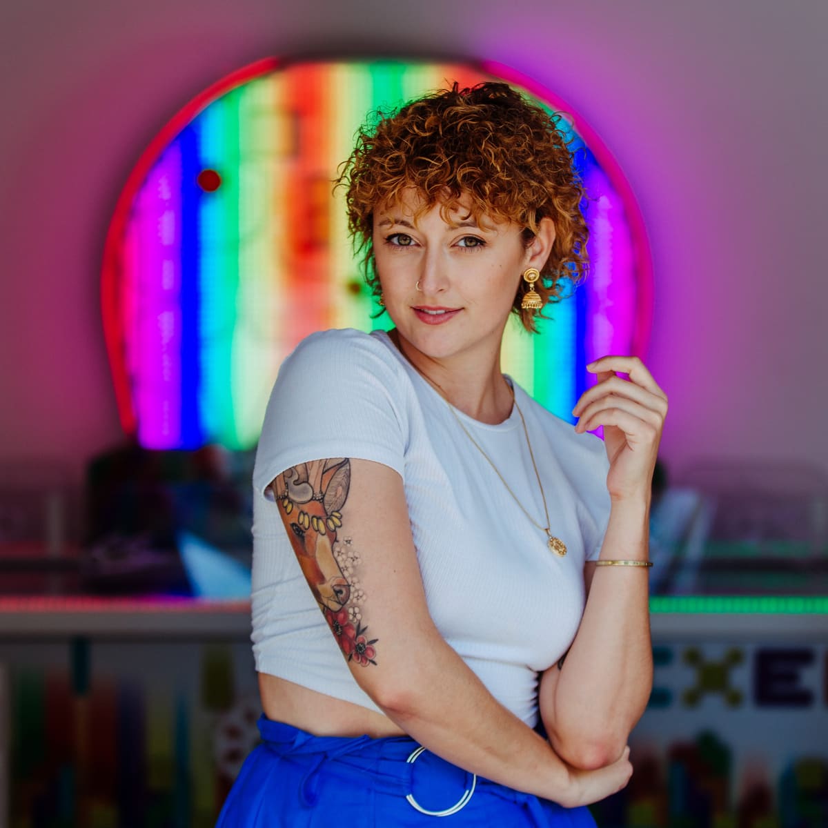 Personal-Branding-Foto einer Frau mit Locken und Tattoo vor einem Regenbogen aus Neonlicht