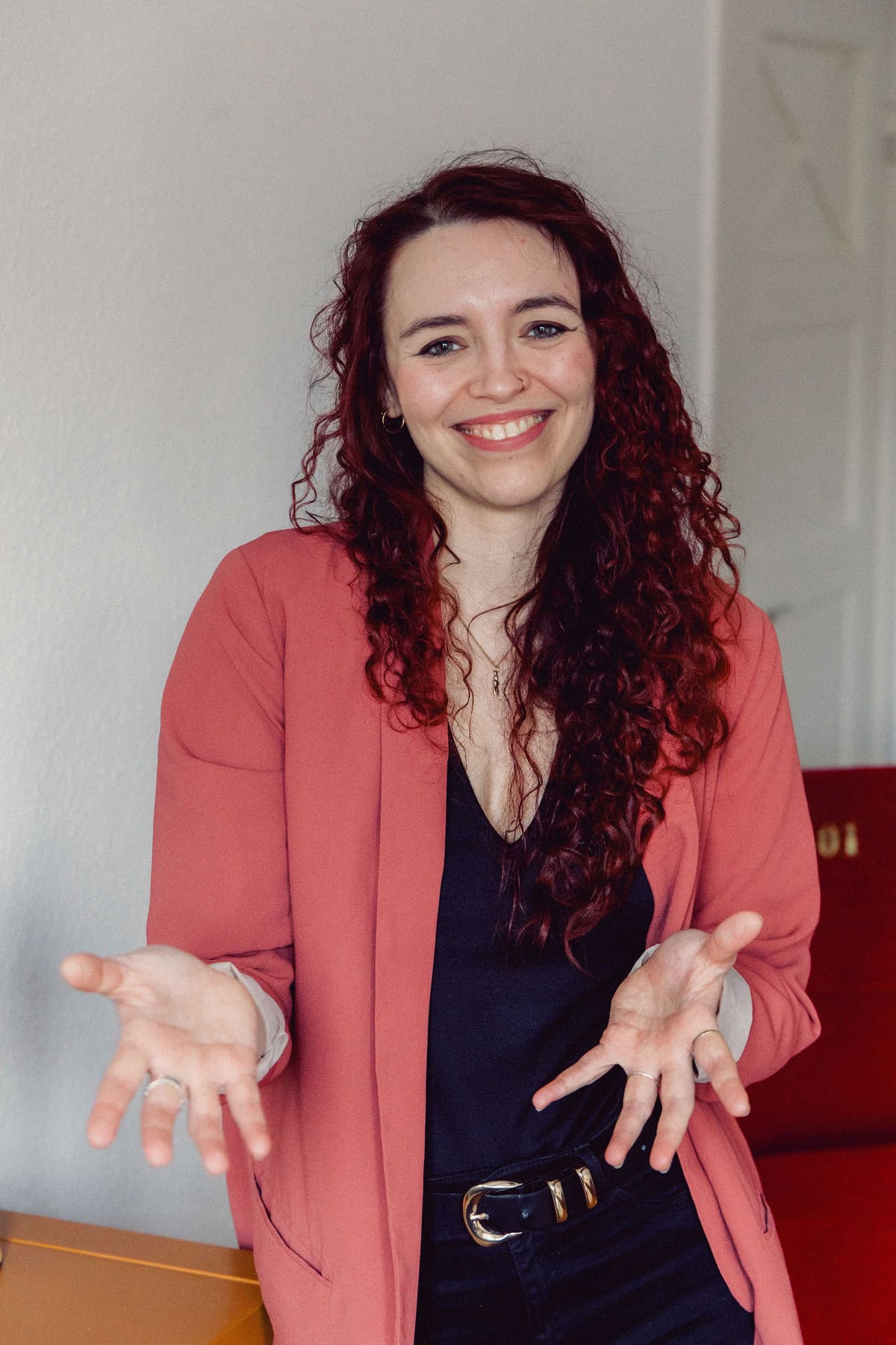 Frau mit roten Locken im korallenfarbenen Blazer lacht und zeigt mit offenen Händen nach vorn