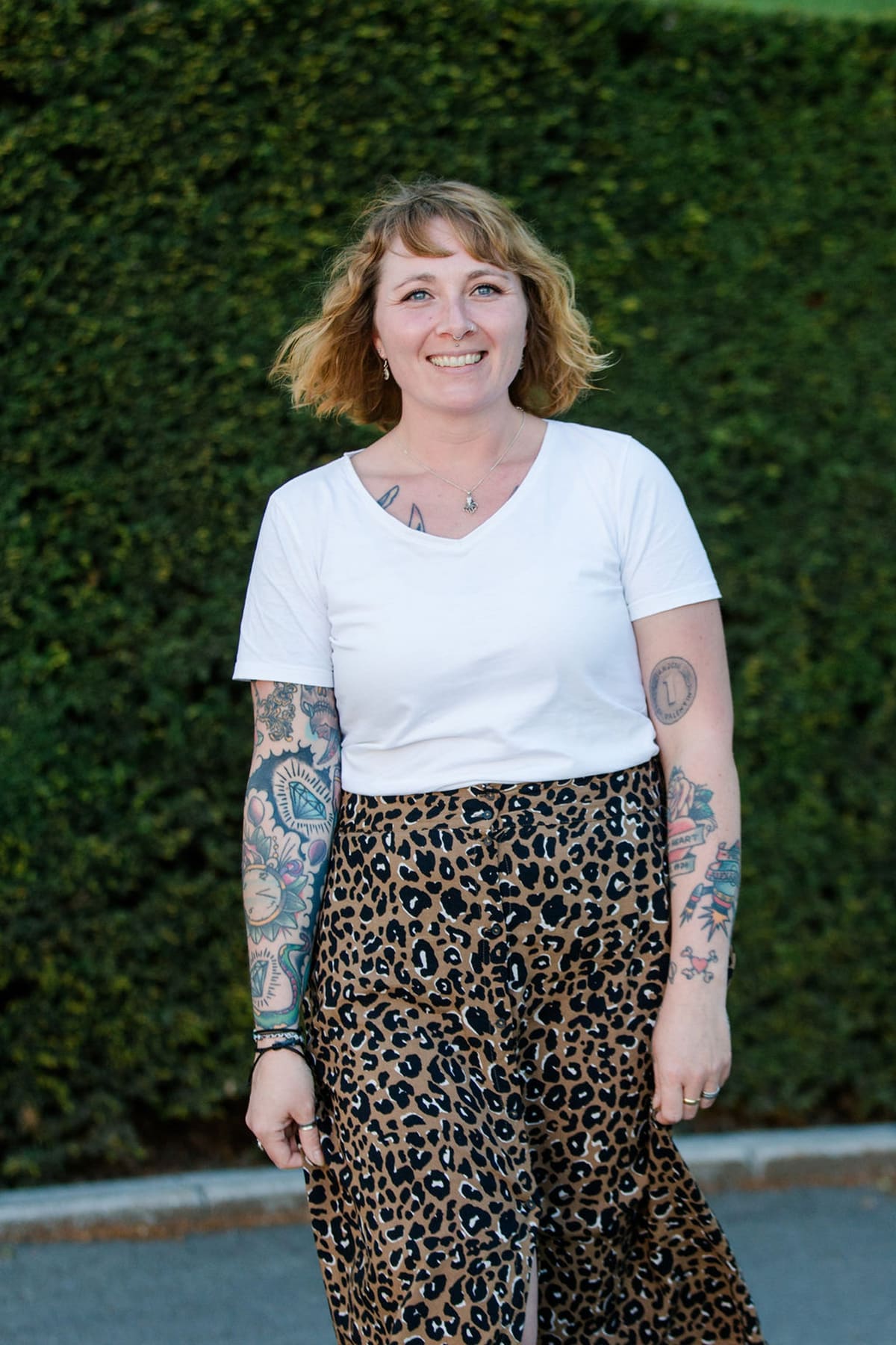 Frau mit Tattoos im weißen Shirt und Leopardenrock vor einer Hecke