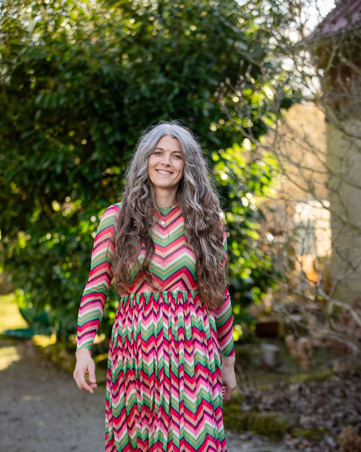 Personal Branding im Garten Frau mit langem grauem Haar im bunten Zickzackkleid steht lächelnd in einem Garten