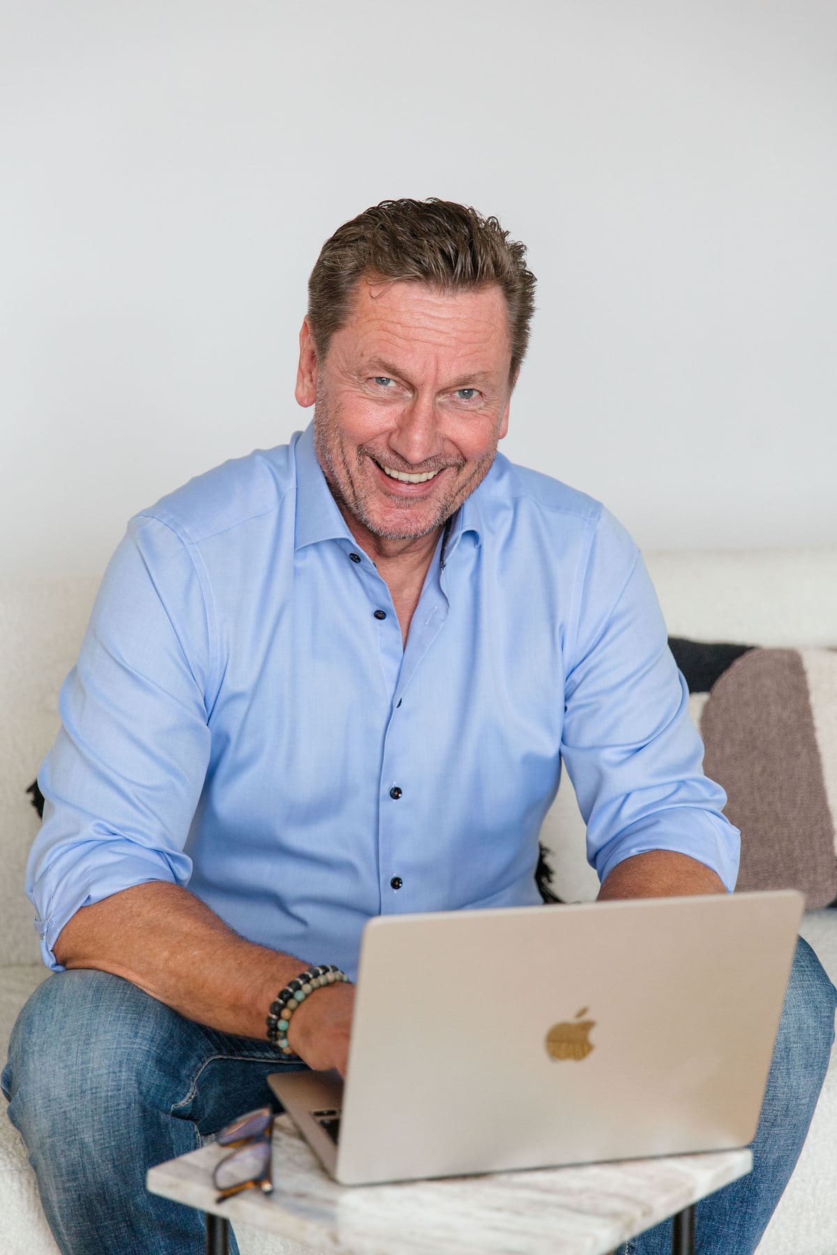 Mann im hellblauen Hemd lacht mit dem Laptop auf dem Sofa