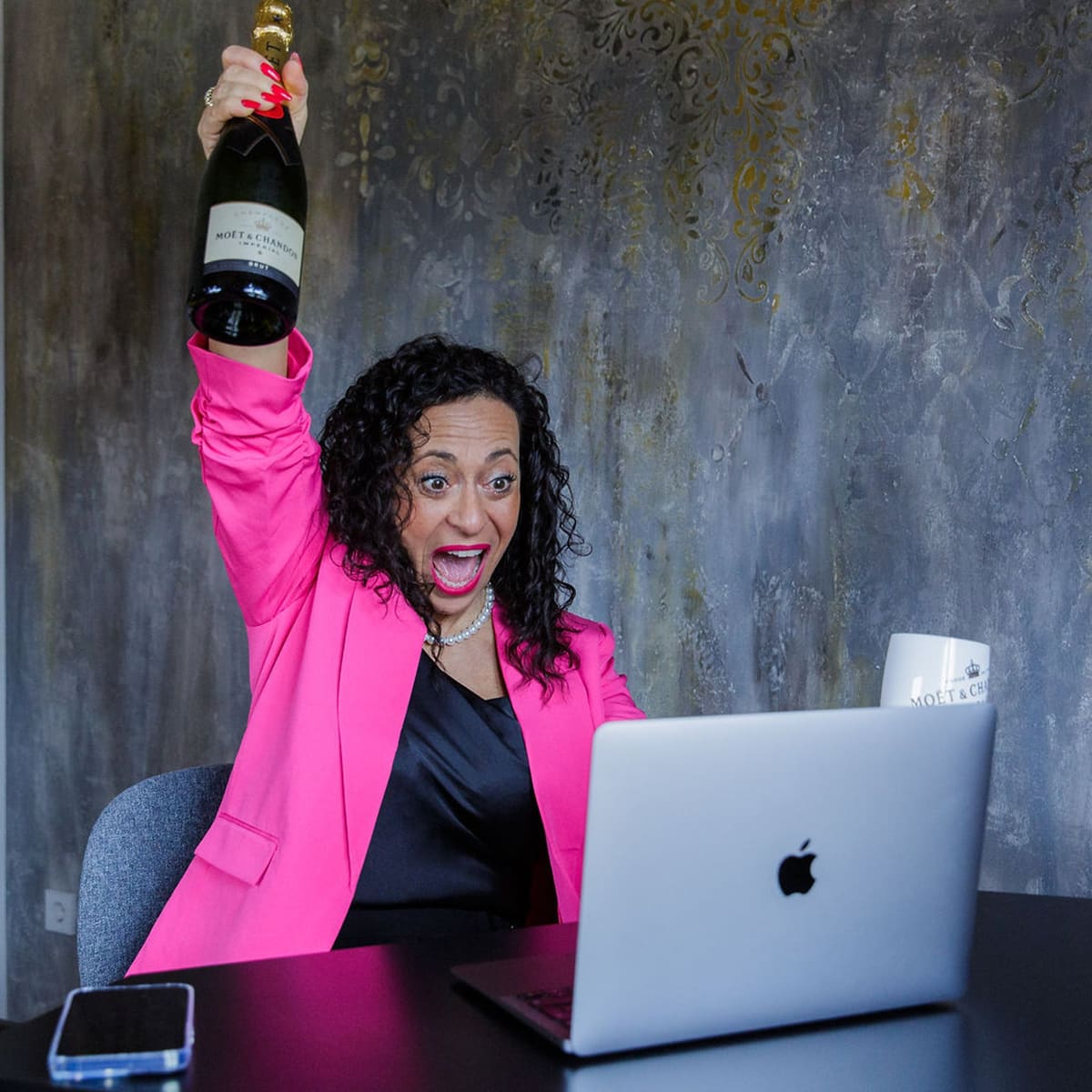 Erfolg feiern am Schreibtisch Unternehmerin im pinken Blazer jubelt am Laptop und hält eine Sektflasche hoch