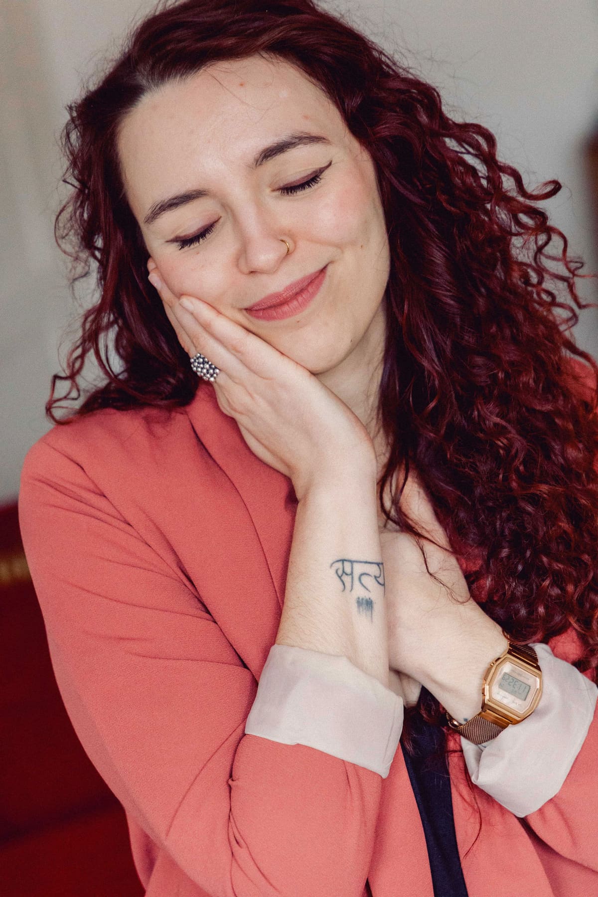 Portrait mit Nähe und Wärme Frau mit roten Locken und korallfarbenem Blazer lächelt mit geschlossenen Augen, die Wange in die Hand gelegt