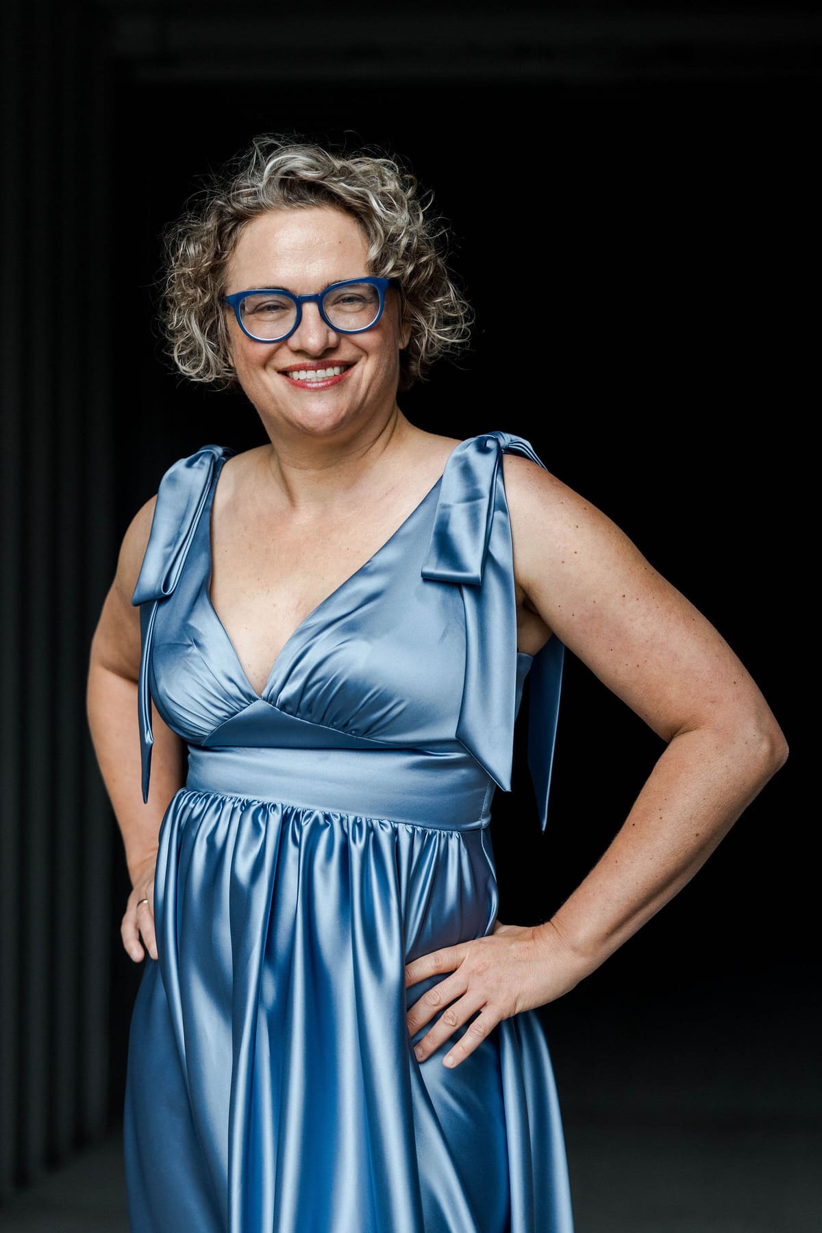 Frau im glänzenden blauen Kleid steht lachend vor dunklem Hintergrund, die Hände an den Hüften